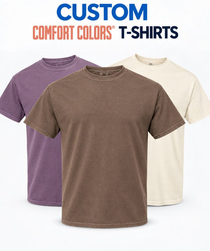 Three short-sleeve crew-neck t-shirts in purple, brown, and cream are displayed against a white background with the text 'CUSTOM COMFORT COLORS T-SHIRTS' at the top.